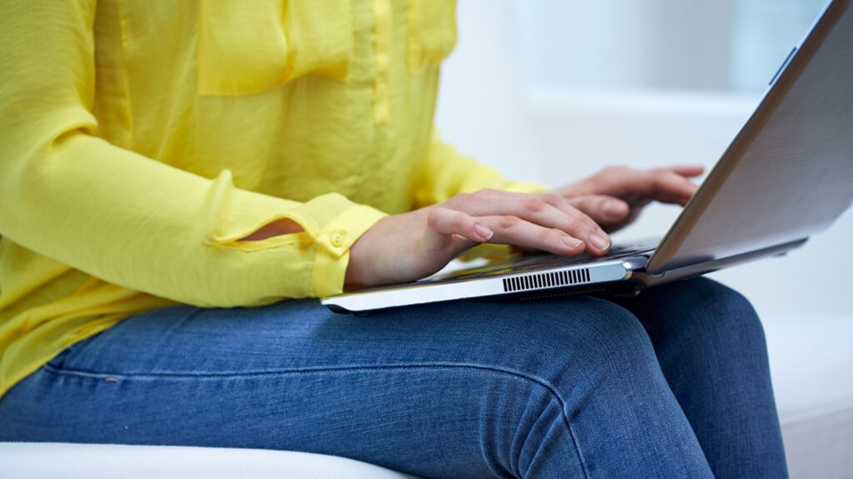 Detaljbild på kvinna i soffa som sitter med uppslagen laptop i en soffa och skriver.