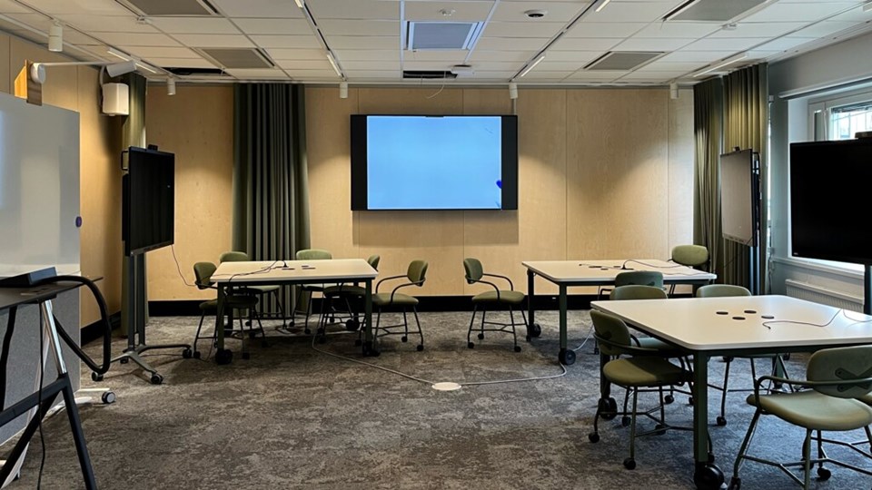 En bild av en tom lärosal med flera skärmar och tre bord med stolar runtom