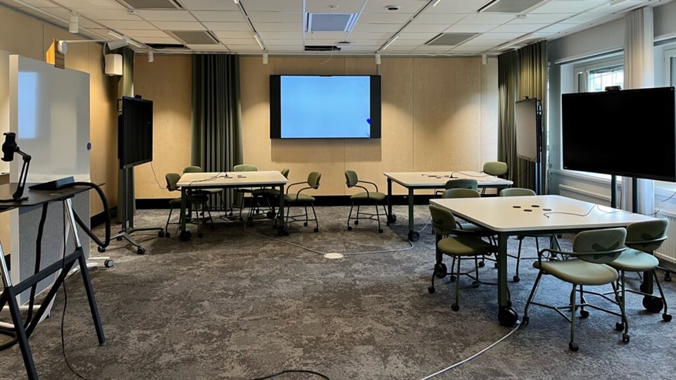 Bilden visar en stor bildskärm, två stora bord med stolar omkring. Ett tomt Learning Lab Umeå