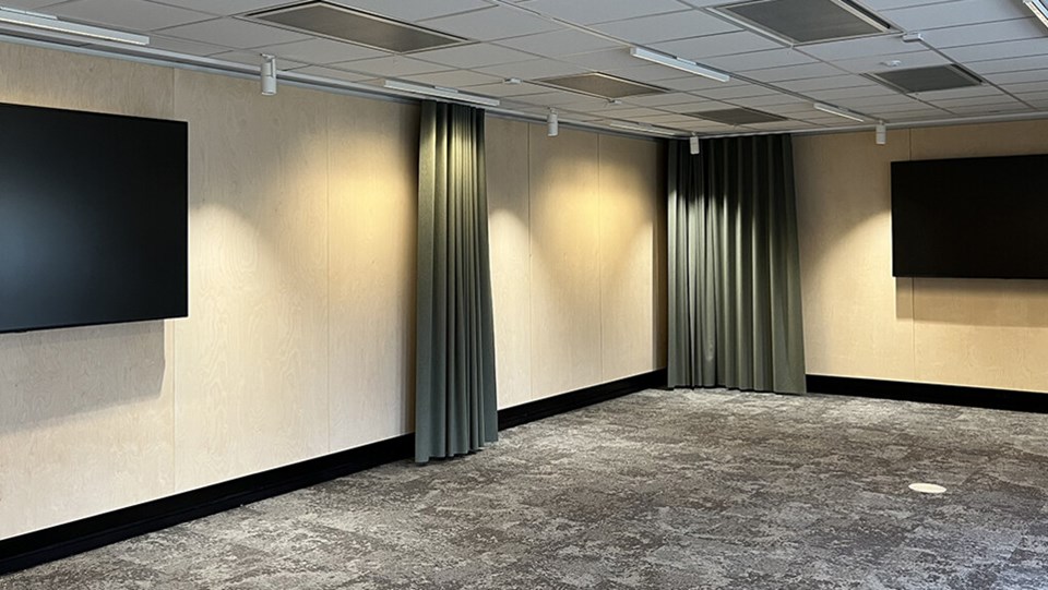 En bild av ett hörn i en lärosal, med gröna draperier och två stora skärmar på väggarna. Golvet är grått och väggarna är i trä.
