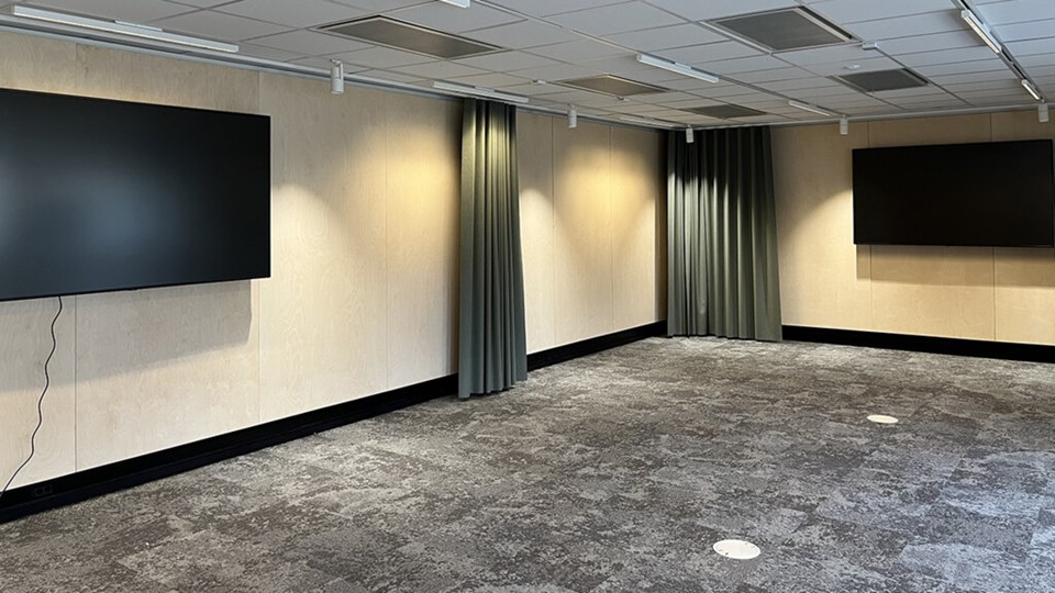 En bild av ett hörn i en lärosal, med gröna draperier och två stora skärmar på väggarna. Golvet är grått och väggarna är i trä.